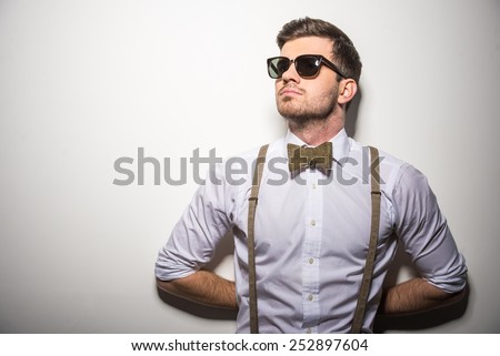 Portrait of young trendy man with black glasses, suspenders and bow-tie on gray background.