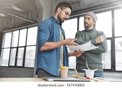 Portrait of young unexperienced male gets consultation from male professional, points at information he doesn`t understand, asks how he can change figures or what should be written, have dinner break