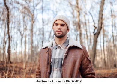 A portrait of a young white man posing in the woods dressed in autumn clothes: brown jacket, checkered canadian style shirt, off-white beanie, looking at the lens