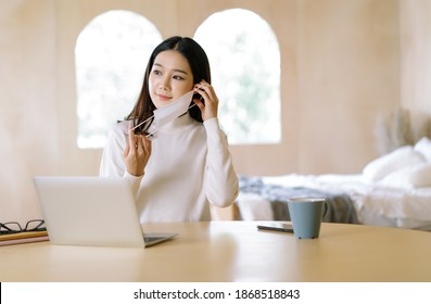 Portrait Of Young Woman Asian Freelancer In Warm Sweater Working On Laptop Prepare To Wear Medical Mask For Protection Flu And Covid-19 At Home.Work On Winter Holidays During Self-isolation Quarantine