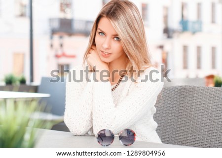 Portrait of a young woman with beautiful blue eyes with a smile with blond hair with natural make-up in a vintage sweater in a summer cafe on the background of  buildings. Attractive girl.