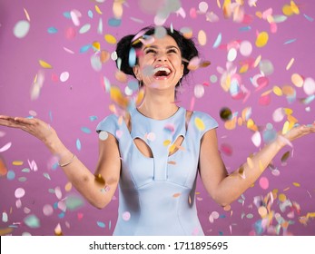 Portrait Of Young Woman With Black Curly Hair In Blue Party Outfit Laughing And Celebrating Confetti Rain Isolated On Pink Background