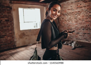 Portrait Of A Young Woman In Black Sportswear Listening To Music From Smart Phone In Hands And Carrying A Sports Bag. Fitness Woman At Gym Looking At Camera And Smiling.