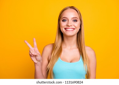 Portrait of young woman with blond hair, big blue eyes and toothy beaming smile. Positive girl demonstrating two fingers v-sign isolated on yellow background with copy space