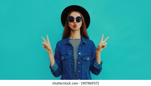 Portrait of young woman blowing her red lips sending air kiss wearing a black round hat, denim jacket on a blue background