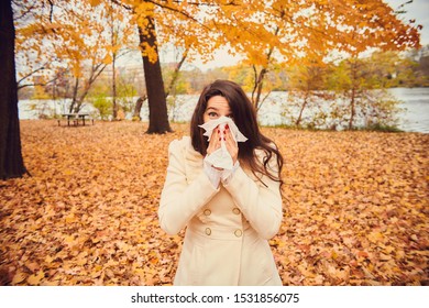 Portrait of young woman blowing nose in autumn park. Girl holding tissue, sneezing or coughing. Cold, flu, allergy