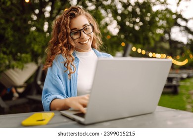 Portrait of a young woman in a blue shirt with a laptop at a table in an outdoor cafe near a lake. Curly woman freelancer works on a laptop outdoors. Freelance concept, nature. - Powered by Shutterstock