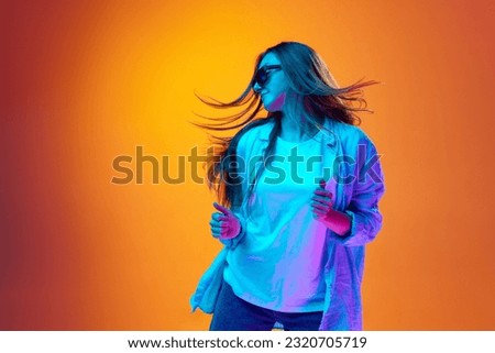 Portrait of young woman in casual clothes and sunglasses dancing against gradient orange studio background in neon light. Concept of youth, human emotions, lifestyle, facial expression, ad