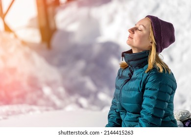 Portrait Of Young Woman With Closed Eyes With Her Head Raised To The Sky And The Sun At The Ski Resort On The Background Of Snow Drifts In The Mountains