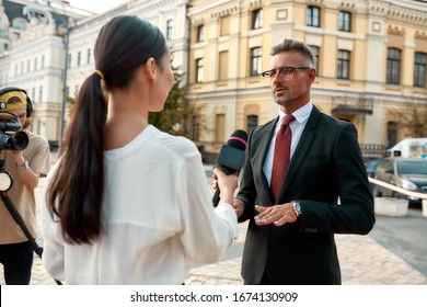 Portrait Of Young Woman Conducting Journalistic Interview Of Cheerful Politician. People Making Interview Using Equipment Set At Outdoor Location. Horizontal Shot. Selective Focus On Man