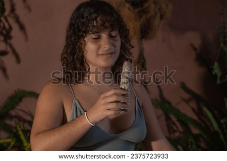 Portrait of a young woman with curly dark hair, sitting crossed legged in meditation holding a smoking smudge stick close to her face