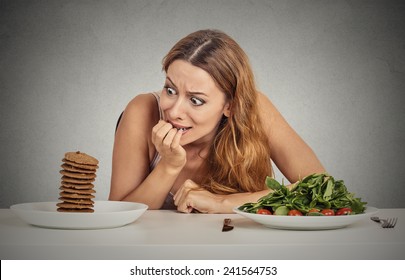 Portrait Young Woman Deciding Whether To Eat Healthy Food Or Sweet Cookies She Is Craving Sitting At Table Isolated Grey Wall Background. Human Face Expression Emotion Reaction Diet Nutrition Concept 