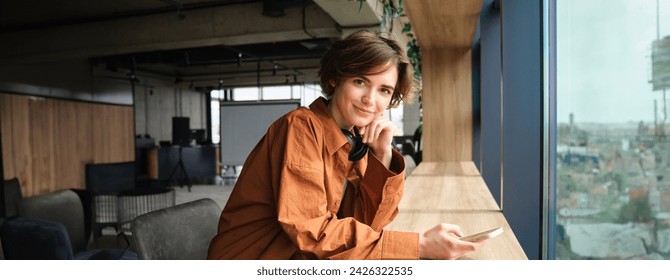 Portrait of young woman, digital nomad working at co-working space, sitting with smartphone, smiling and looking at camera. - Powered by Shutterstock