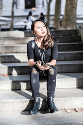 Portrait Of Young Woman Dressed In Black Sitting On Steps Listening Music With Earphones And Cell Phone
