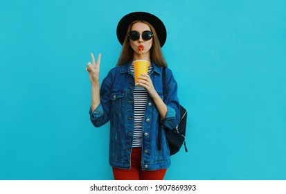 Portrait of young woman drinking a juice wearing a black round hat, denim jacket on a blue background