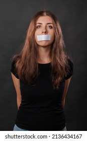 Portrait of a young woman with duct tape sealed in her mouth, restriction of freedom of speech and censored and forbidden to speak and express her opinion, isolated on a dark background.
				