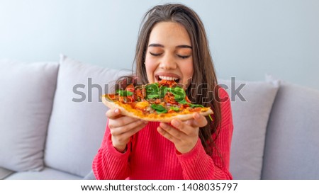 Portrait of a young woman eating pizza, her eyes closed with pleasure, sitting at the living room. Funny brunette girl in red sweater eating pizza. Happy woman eating tasty pizza. 
