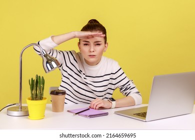 Portrait Of Young Woman Employee Sitting At Workplace With Laptop And Looking Far Away With Hand Above Eyes, Searching With View. Indoor Studio Studio Shot Isolated On Yellow Background.