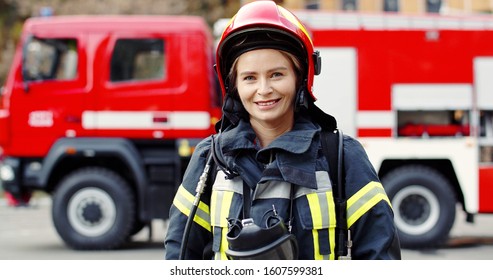 Portrait of young woman firefighter standing near fire truck. Fireman in protective suit with oxygen mask and helmet. 