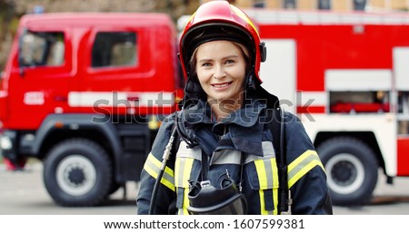 Portrait of young woman firefighter standing near fire truck. Fireman in protective suit with oxygen mask and helmet. 