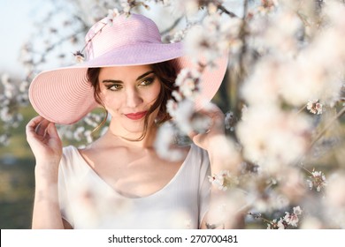 Portrait Of Young Woman In The Flowered Field In The Spring Time. Almond Flowers Blossoms. Girl Wearing White Dress And Pink Sun Hat