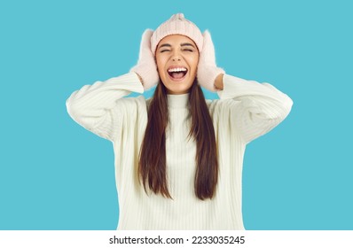 Portrait of young woman frantically reacting to huge winter sales and crazy holiday discounts. Caucasian woman in mittens and hat grabs head and screams excitedly isolated on light blue background.