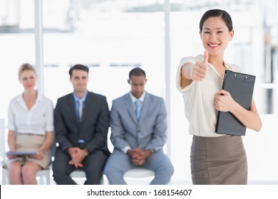 Portrait of a young woman gesturing thumbs up with people waiting for job interview in a bright office - Powered by Shutterstock