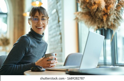 Portrait young woman in glasses drinking coffee, smiling to camera, enjoying studying in coworking space. female sitting in front open laptop computer. Study, learning, remote work, freelance.