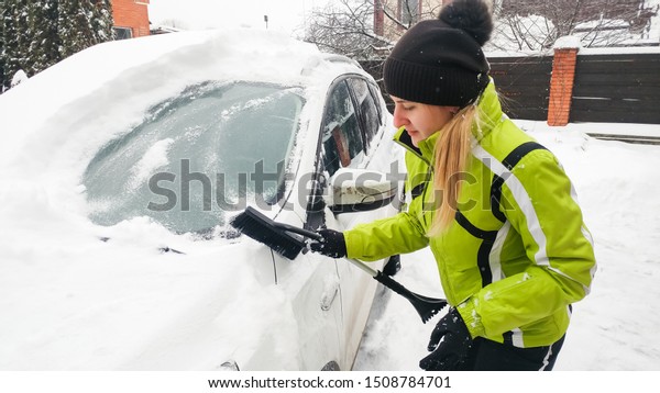Portrait of young woman in\
green coat is getting ready to clean snow off a white car using a\
snow brush.