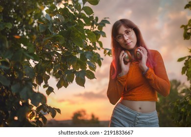 Portrait Of A Young Woman With Headphones In Her Hands Looking Ahead At Sunset In A  Park.