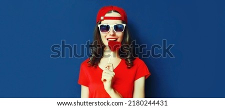Portrait of young woman with heart shaped red lollipop wearing baseball cap, sunglasses on blue background