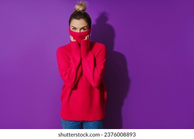 Portrait of young woman is hiding her face inside a warm and cozy polo neck sweater against purple background - Shutterstock ID 2376008893