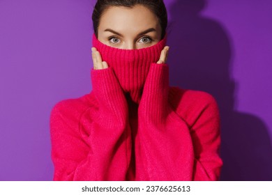 Portrait of young woman is hiding her face inside a warm and cozy polo neck sweater against purple background - Shutterstock ID 2376625613