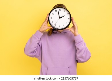 Portrait Of Young Woman Holding Wall Clock Hiding Her Face, Time Management, Schedule And Meeting Appointment, Wearing Purple Hoodie. Indoor Studio Shot Isolated On Yellow Background.