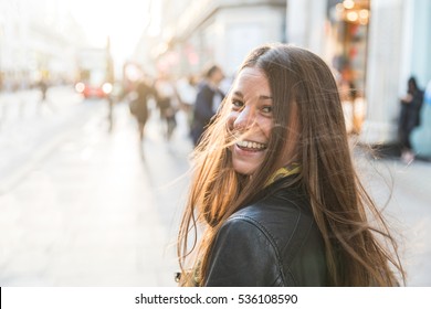 Portrait Of A Young Woman In London. Smiling Girl Looking Away From Camera. She Is Standing On The Pavement Next To The Street, With Blurred People And Traffic On Background.