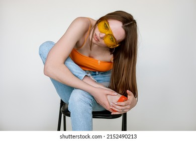 Portrait of young woman with long dark hair wearing orange top, blue jeans, sunglasses, sitting on chair, bending down.
