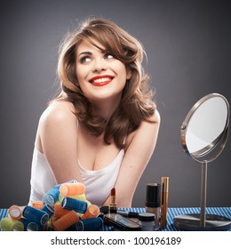 Portrait Of A Young Woman With Long Hair On Gray Background Isolated.  Happy Girl Seating At Table With Make Up Accessories And Mirror. Smiling Model With Curler Hair Dress