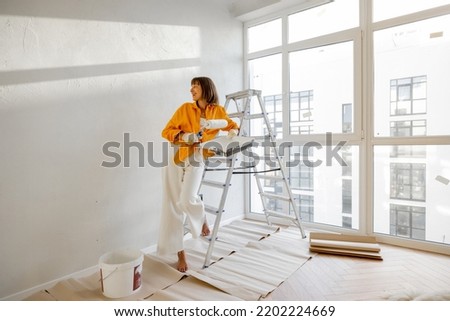 Portrait of a young woman making repairing in apartment, standing with paint roller near ladder in bright room of her new home