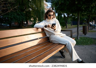 Portrait of a young woman in a modern sun glasses sitting on the wooden bench in a park near a lake enjoying the sunset.Stylish girl in light sports clothes sitting and relaxing on the beach at sunset - Powered by Shutterstock