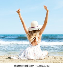 Portrait Of Young Woman On The Beach Near The Sea Sitting With Hands Up Wearing Sophistical Dress And Hat. Photo From Behind