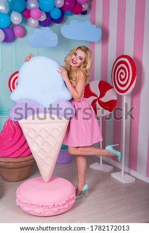 Portrait of young woman in pink dress holding big ice cream and posing on decorated background. Amazing sweet-tooth lady surrounded by toy sweets. 