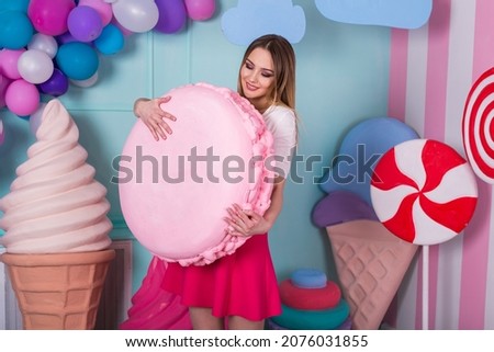 Portrait of young woman in pink dress holding big macaroon and posing on decorated background. Amazing sweet-tooth girl surrounded by toy sweets. 