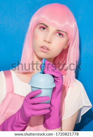 Portrait of young woman with pink hair drinking from disposable blue plastic cup. Teenager holding summer cocktail in protective gloves on blue background. Advertising protection during coronavirus.