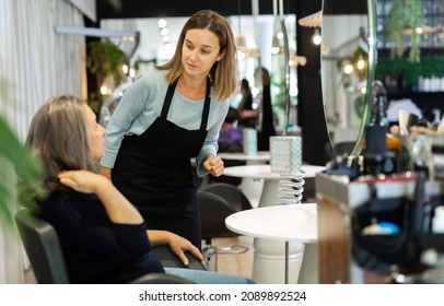 Portrait Of Young Woman Professional Hair Stylist Talking To Elderly Female Client In Salon, Choosing New Hairdo .