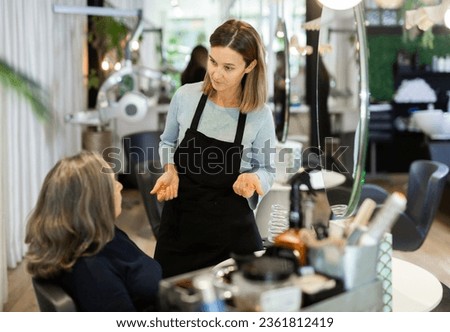 Portrait of young woman professional hair stylist talking to elderly female client in salon, choosing new hairdo .