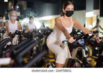 Portrait Of Young Woman In Protective Face Mask Training On Stationary Bike Workout In Gym