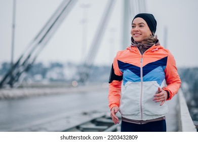 Portrait Of A Young Woman Running And Training On A Bridge On A Cold Winter Day. Happy Athletic Woman Jogging Outside At Winter. Healthy Lifestyle. Wearing An Active Wear And A Black Cap. Copy Space.