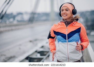 Portrait Of A Young Woman Running And Training On A Bridge On A Cold Winter Day. Happy Athletic Woman Jogging Outside At Winter. Copy Space. Healthy Lifestyle. Using Headphones And Listening Music.