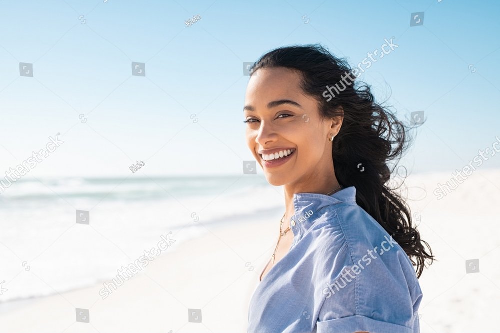 Portrait of young woman at sea looking at camera. Smiling latin hispanic girl standing at the beach with copy space and looking at camera. Happy mixed race girl in casual outfit with wind in her hair.