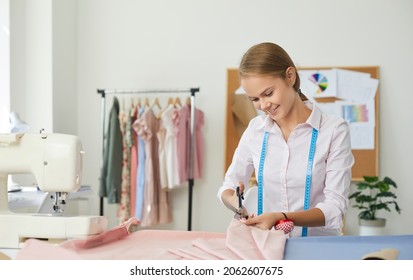 Portrait Of Young Woman Seamstress Who Uses Scissors To Cut Out Detail Of Dress On Lines Of Sketch. Friendly Smiling Woman Working In Her Own Sewing Studio. Concept Of Small Business.